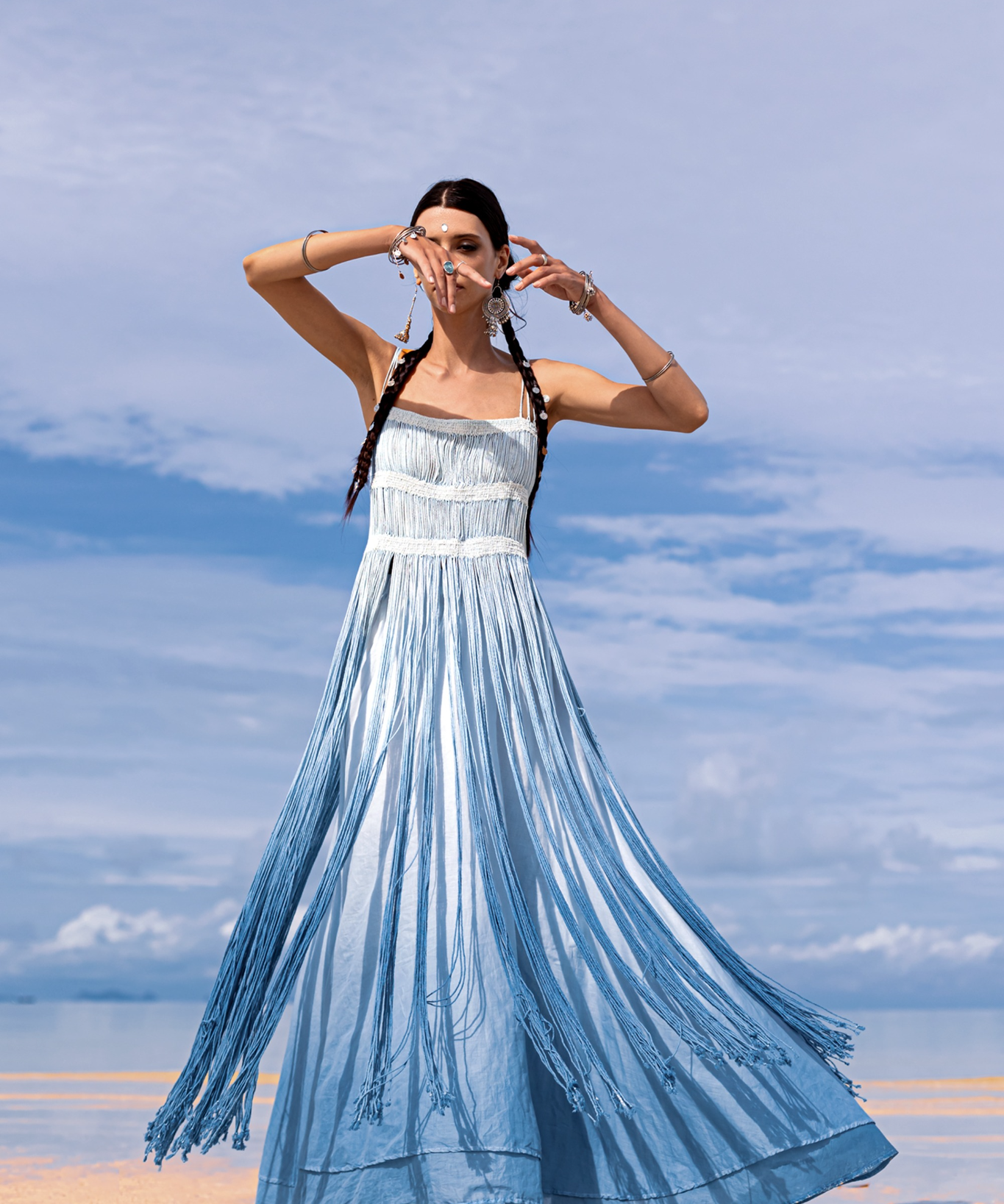 A woman stands barefoot on a sandy beach, wearing an AYA Sacred Wear Sky Blue Greek Goddess Dress botanically dyed in blue shades of organic cotton. Her braided hair and tribal bracelets sway in the gentle breeze, under a sky dotted with clouds as the ocean softly whispers nearby.