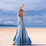 A woman stands on a sandy beach, arms gracefully raised, wearing the AYA Sacred Wear Sky Blue Greek Goddess Dress—a boho, botanically dyed creation made from organic cotton. Framed by ocean and distant hills under a sky with scattered clouds, she embodies elegance.