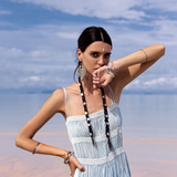 On the beach, a woman in a Sky Blue Greek Goddess Dress by AYA Sacred Wear stands with two braided hair adorned with beads. Her arm showcases silver bracelets against the peaceful beach and cloudy sky.