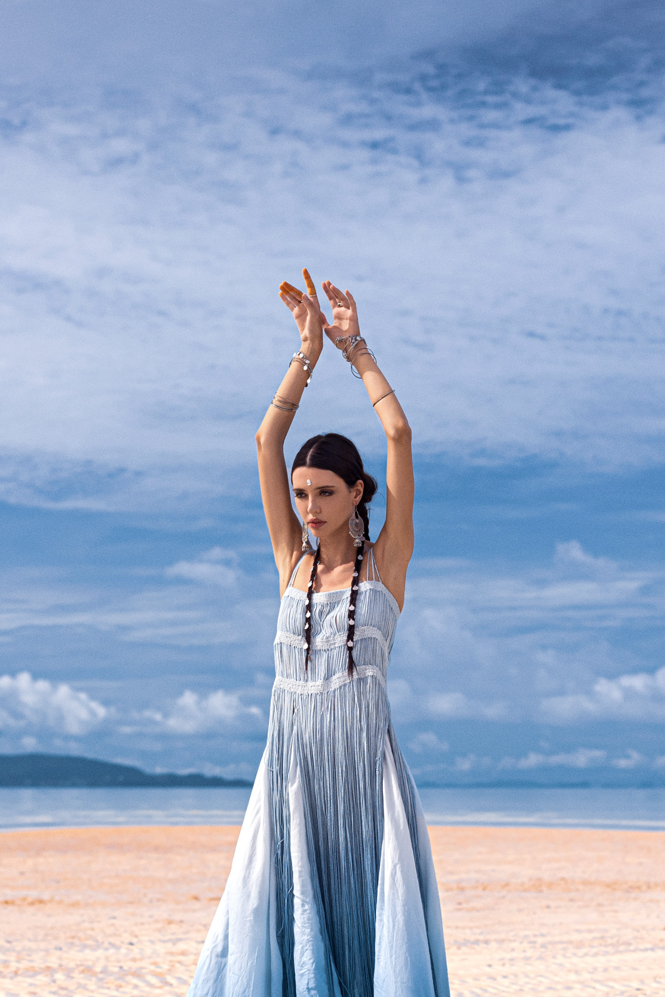 A woman in an AYA Sacred Wear Sky Blue Greek Goddess Dress stands on a beach with raised arms. The partly cloudy sky and ocean backdrop highlight her tribal-themed outfit, accented by long earrings and bracelets catching the coastal breeze.