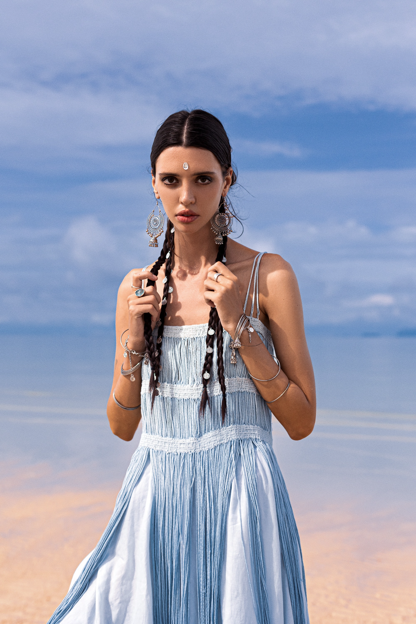A woman with long braided hair stands on a sandy beach wearing AYA Sacred Wear's Sky Blue Greek Goddess Dress, sleeveless and crafted from organic cotton. Intricate earrings and bracelets enhance her look as the dress's botanically dyed fabric complements the sea and sky behind her cloudy backdrop.