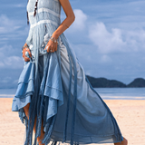 Barefoot on a sandy beach, a person wears AYA Sacred Wear's Sky Blue Greek Goddess Dress, an ombre macrame piece with spaghetti straps and botanical dyes. It complements their long braids and large earrings under a partly cloudy sky with distant hills visible.