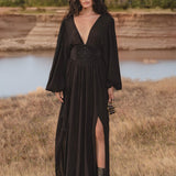 A woman with long wavy hair stands outdoors near water, wearing the AYA Sacred Wear Tatva Dress Black Colour – a handmade, botanically dyed natural cotton linen V front-back kaftan with a high slit, paired with black ankle boots.