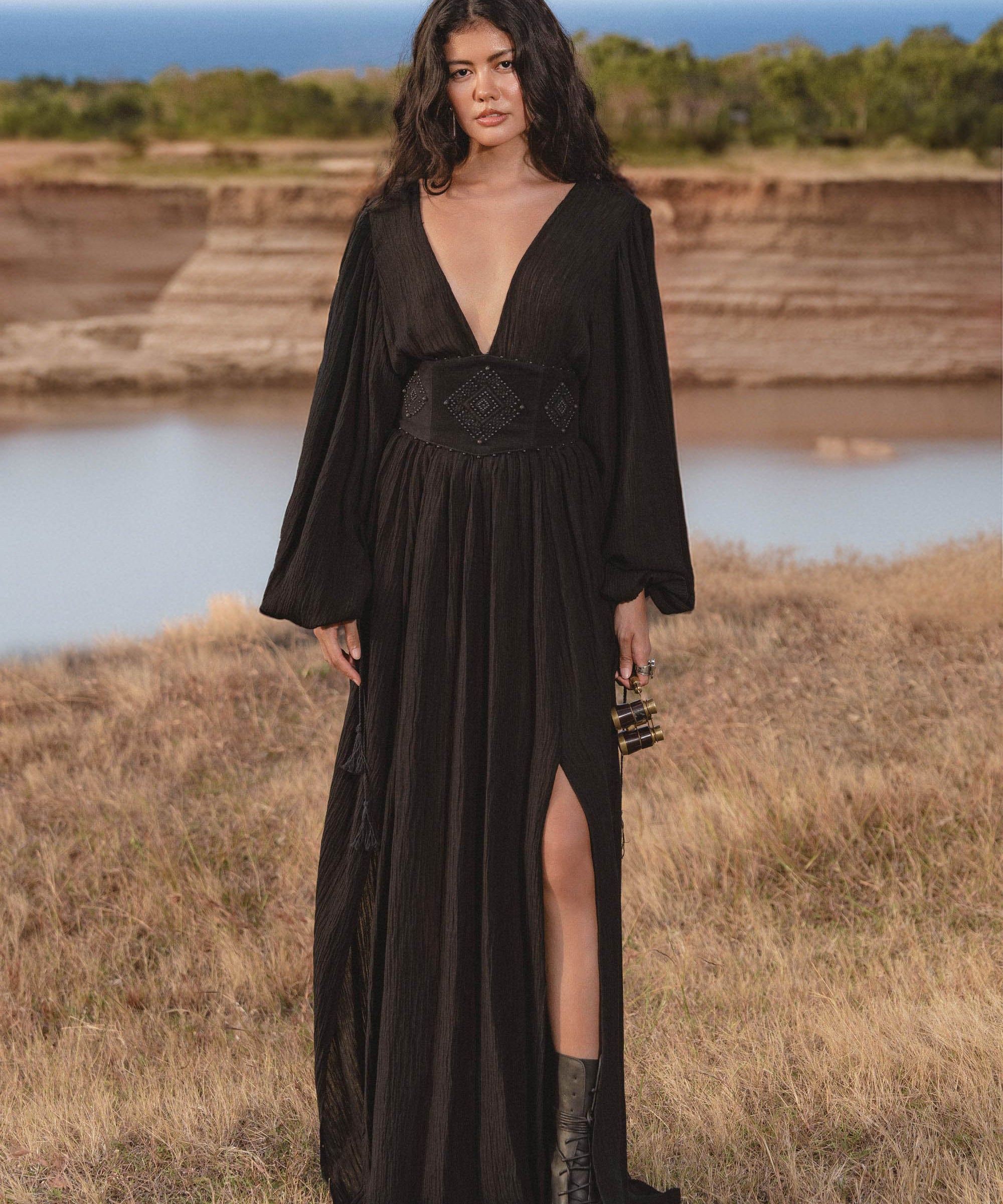 A woman with long wavy hair stands outdoors near water, wearing the AYA Sacred Wear Tatva Dress Black Colour – a handmade, botanically dyed natural cotton linen V front-back kaftan with a high slit, paired with black ankle boots.