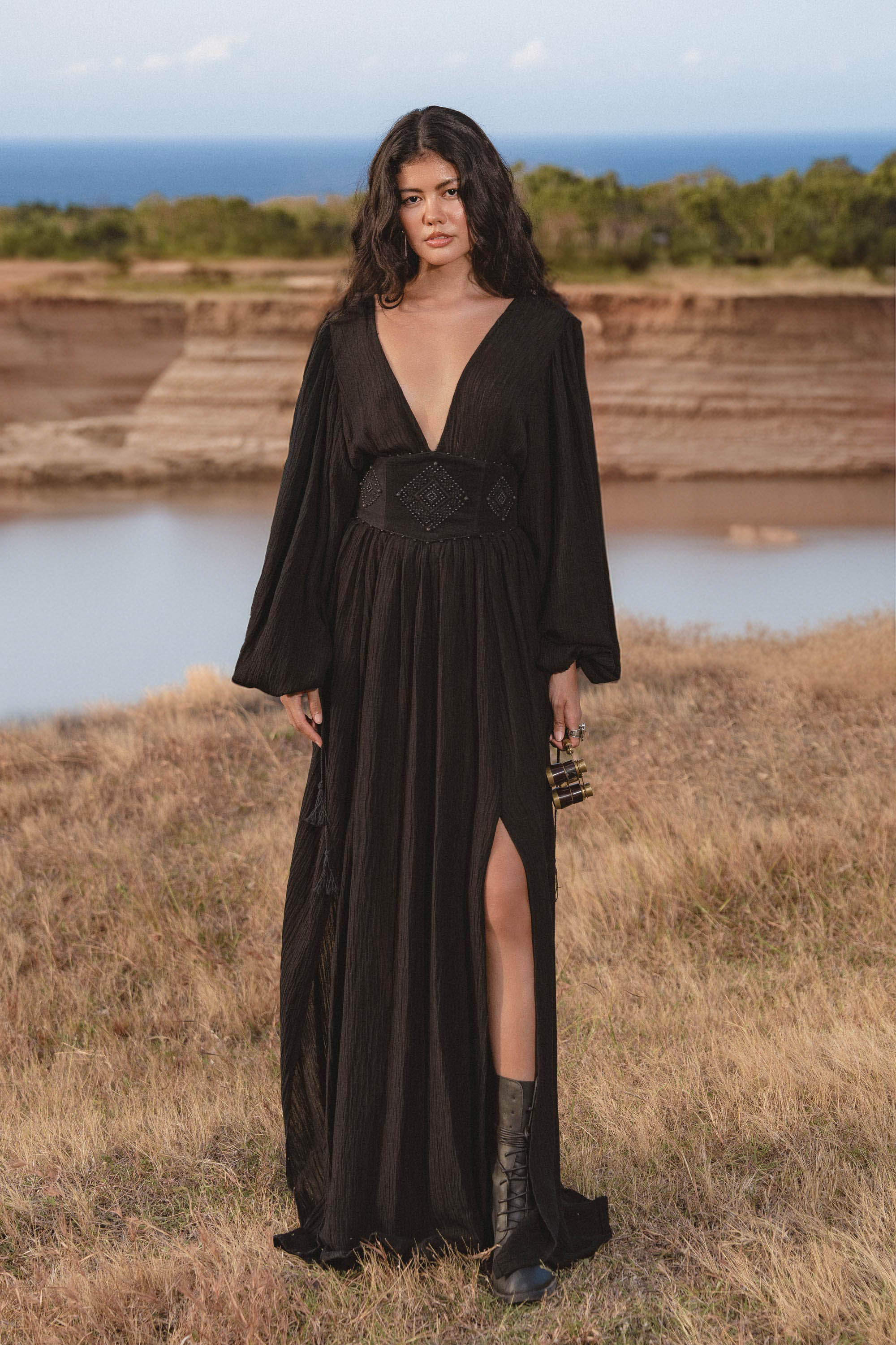 A woman with long wavy hair stands outdoors near water, wearing the AYA Sacred Wear Tatva Dress Black Colour – a handmade, botanically dyed natural cotton linen V front-back kaftan with a high slit, paired with black ankle boots.