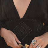 Wearing the AYA Sacred Wear Tatva Dress Black—a handmade V front-back kaftan in natural cotton linen with a wide beaded geometric belt—silver rings adorn their hands as they hold vintage opera glasses; only torso and hands are seen.