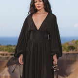A woman with long dark hair wears the Tatva Dress Black Colour – Handmade V Front Back Kaftan by AYA Sacred Wear, standing outdoors before grassy hills and a blue sky, holding a small clutch and looking calmly at the camera.