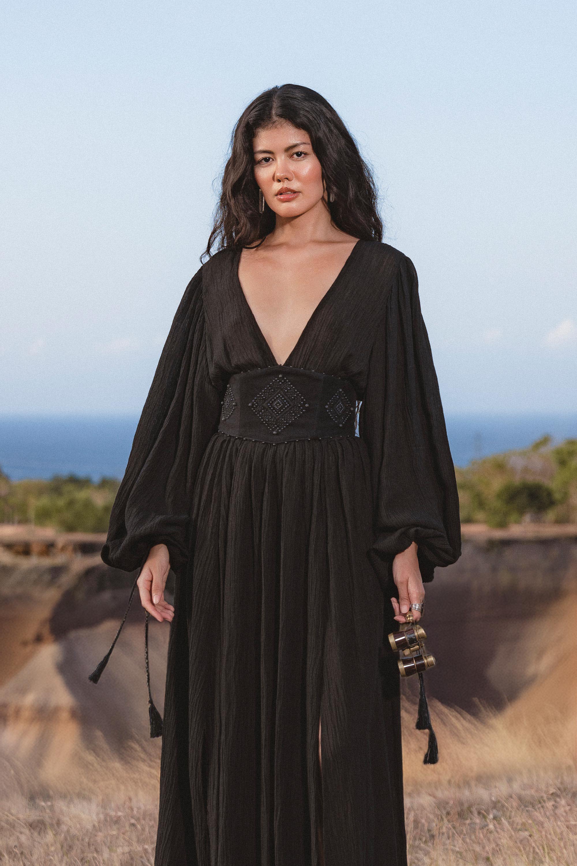 A woman with long dark hair wears the Tatva Dress Black Colour – Handmade V Front Back Kaftan by AYA Sacred Wear, standing outdoors before grassy hills and a blue sky, holding a small clutch and looking calmly at the camera.