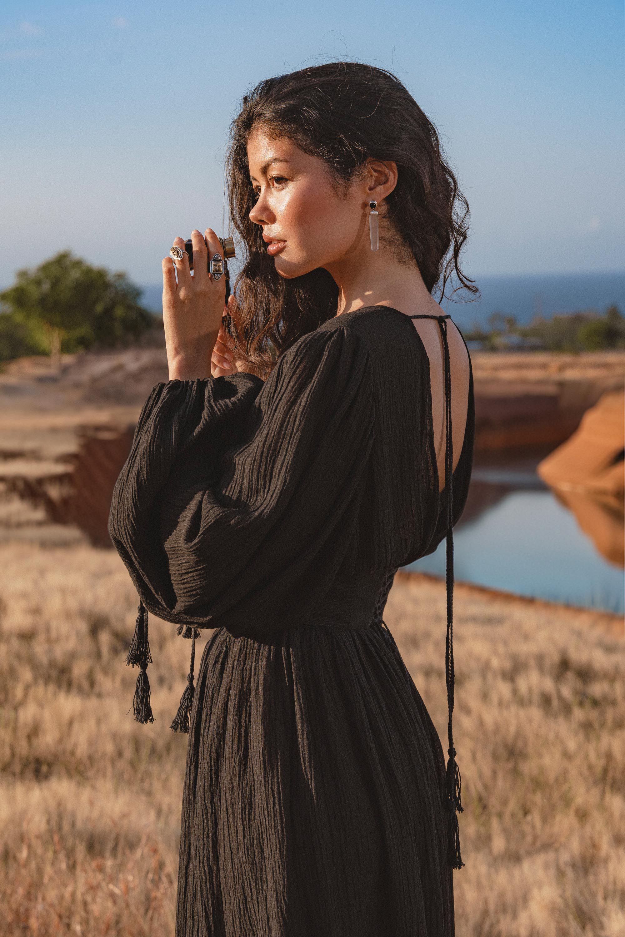 A woman with long wavy hair stands outdoors in profile, wearing the AYA Sacred Wear Tatva Dress Black Colour – a handmade V front-back kaftan crafted from botanical-dyed cotton linen, gazing thoughtfully by a pond under a blue sky.