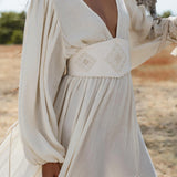 A woman wears the Tatva Dress in Ivory by AYA Sacred Wear—a sustainable bohemian dress with a deep V-neck, high waist, flowy skirt with front slit, and a handcrafted belt, standing outdoors amid dry grass and greenery while holding an owl.
