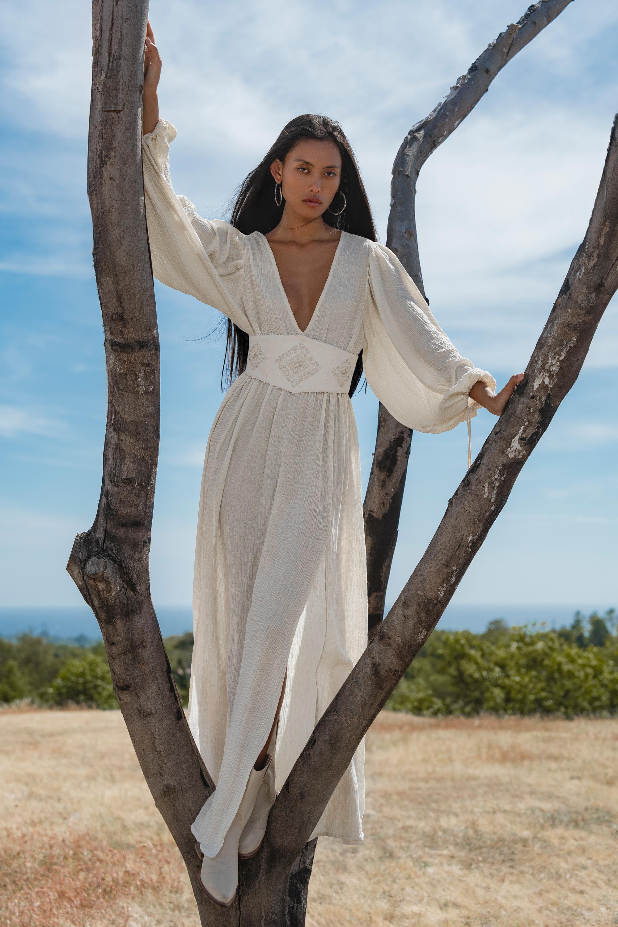 A woman in a flowing, long-sleeved Tatva Dress Ivory Colour – Sustainable Bohemian Dress by AYA Sacred Wear stands between bare tree branches in a grassy field, gazing calmly ahead under a blue sky with distant greenery behind her.
