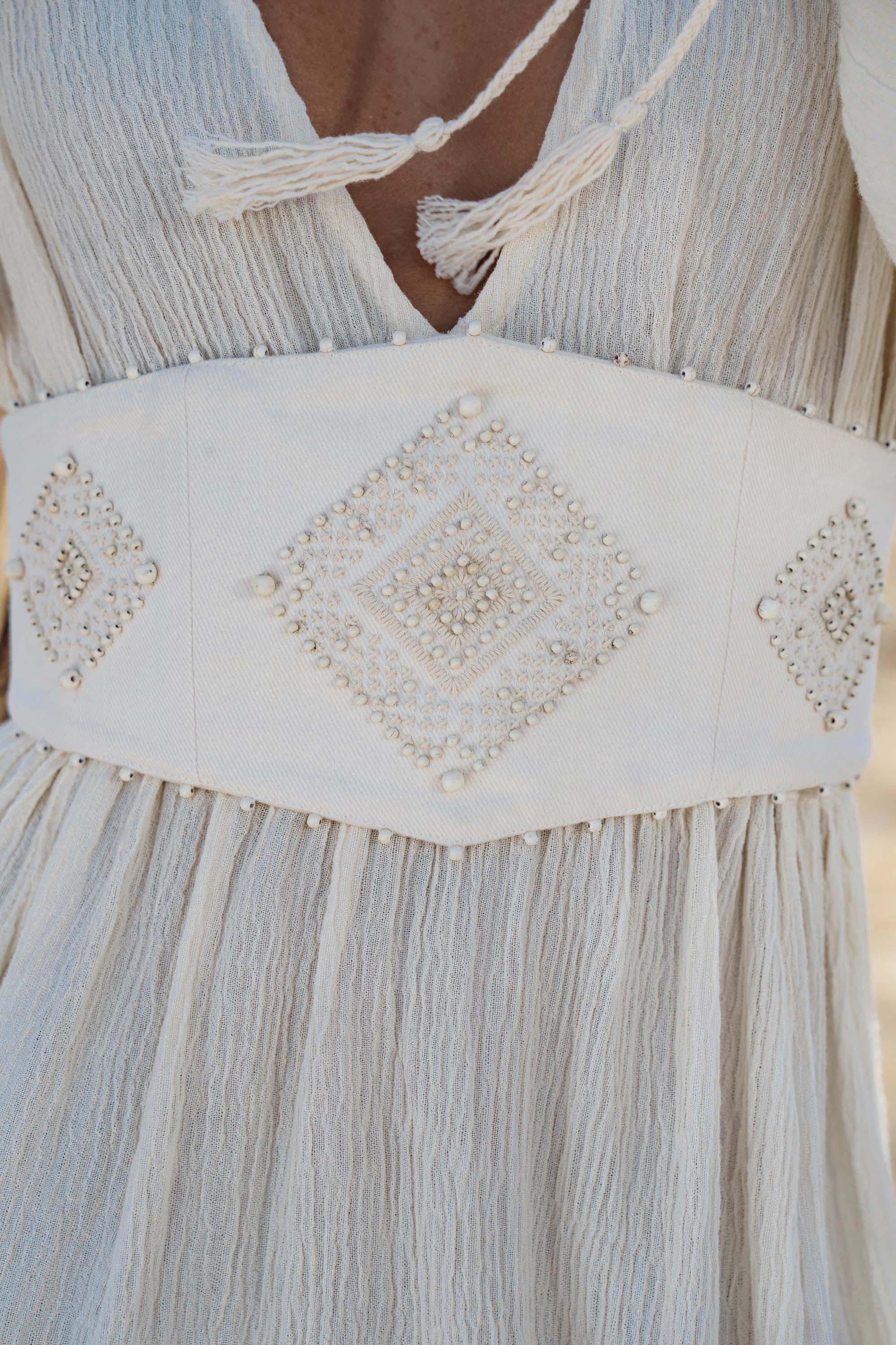 A close-up of the AYA Sacred Wear Tatva Dress Ivory Colour – Sustainable Bohemian Dress highlights its white botanically dyed fabric, wide handcrafted belt with pearl-beaded diamond motifs, and tasseled V-neck tie.
