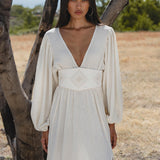 A woman with long dark hair stands outdoors by a tree, wearing the Tatva Dress Ivory Colour—a sustainable, bohemian dress by AYA Sacred Wear with long sleeves, a deep V-neck, and a wide handcrafted embroidered belt. Nature surrounds her.
