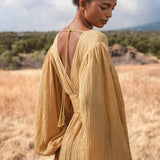 A woman with dark hair in a bun stands in a grassy field, wearing the AYA Sacred Wear Tatva Dress Saffron Gold Colour—an organic, loose-fit dress with long sleeves and deep V-back—framed by trees and a partly cloudy sky.
