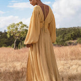 In a dry grassy field, a woman wearing the Tatva Dress Saffron Gold Colour by AYA Sacred Wear stands beneath a partly cloudy sky. The organic, flowing dress features billowy sleeves and an open back, her hair tied as trees frame the serene scene.