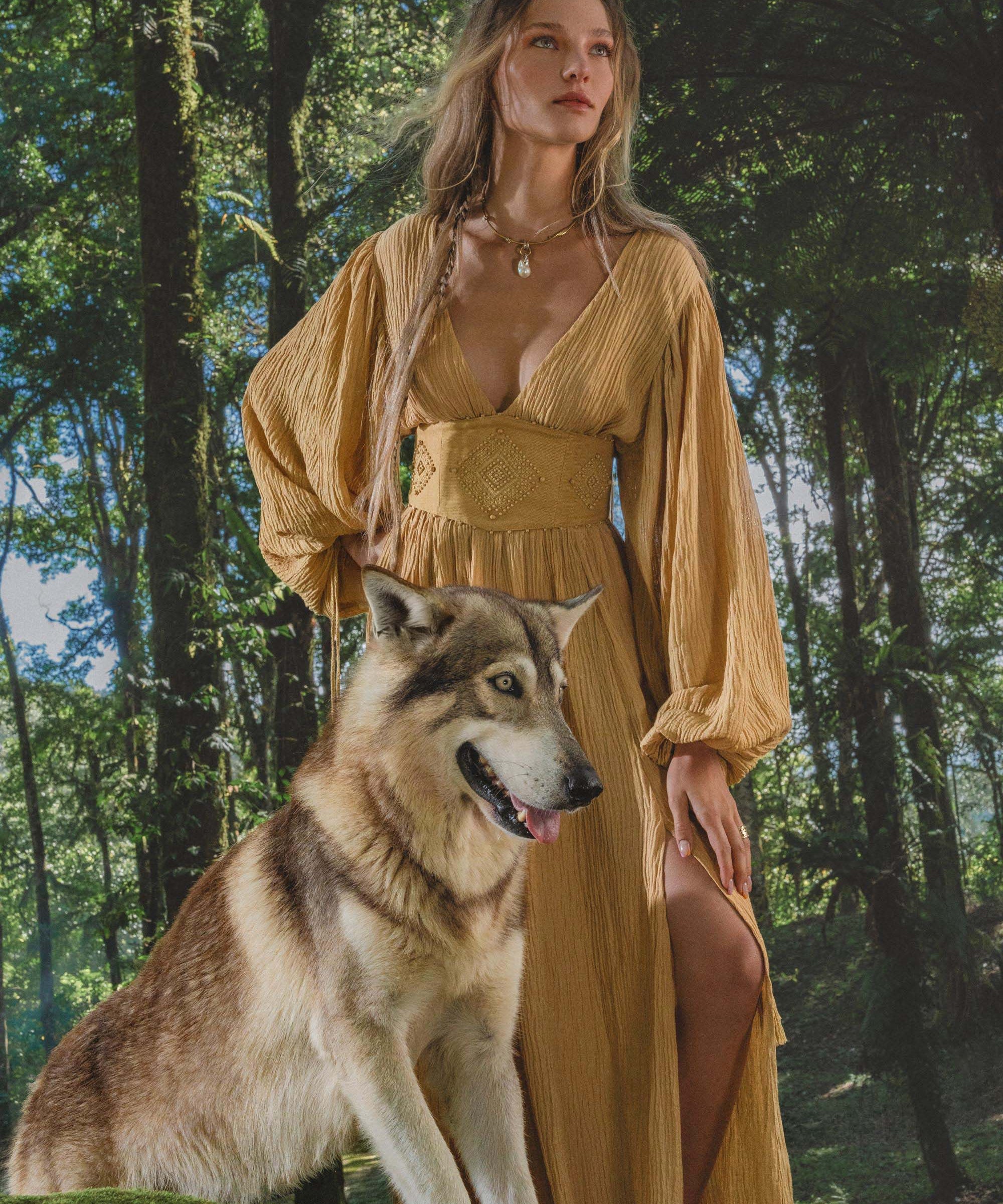 A woman with long wavy hair wears the Tatva Dress Saffron Gold Colour by AYA Sacred Wear, standing calmly in a lush forest with sunlight streaming through the trees and her hand resting on a large, wolf-like dog beside her.
