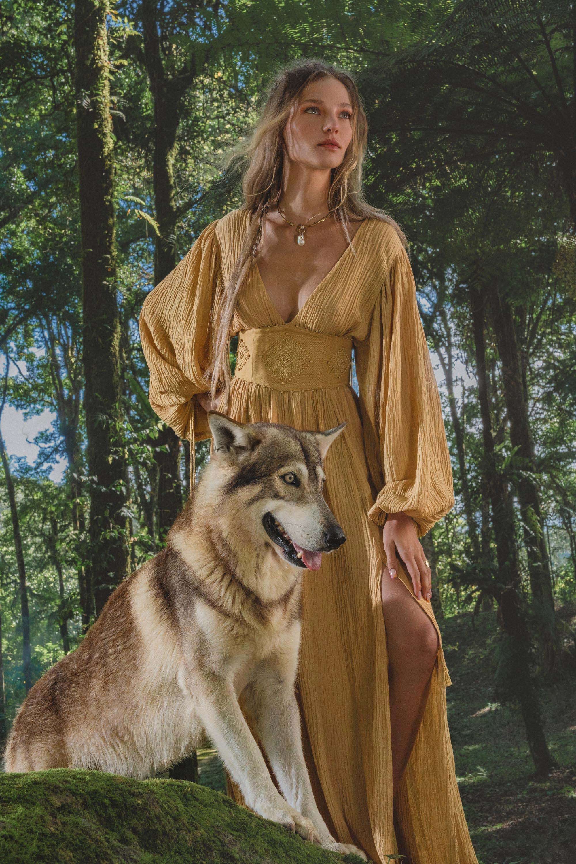 A woman with long wavy hair wears the Tatva Dress Saffron Gold Colour by AYA Sacred Wear, standing calmly in a lush forest with sunlight streaming through the trees and her hand resting on a large, wolf-like dog beside her.