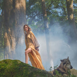 A woman in an AYA Sacred Wear Tatva Dress Saffron Gold Colour – Organic Goddess Dresses stands on a mossy rock in a sunlit forest, mist drifting among the trees as a watchful gray wolf lies beside her under filtered sunlight.
