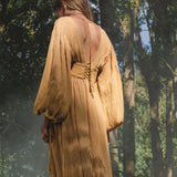 A person stands in a sunlit forest wearing the AYA Sacred Wear Tatva Dress in Saffron Gold—a botanically dyed, organic dress with voluminous sleeves and an open back tied at the neck. Soft light creates a serene, natural setting.