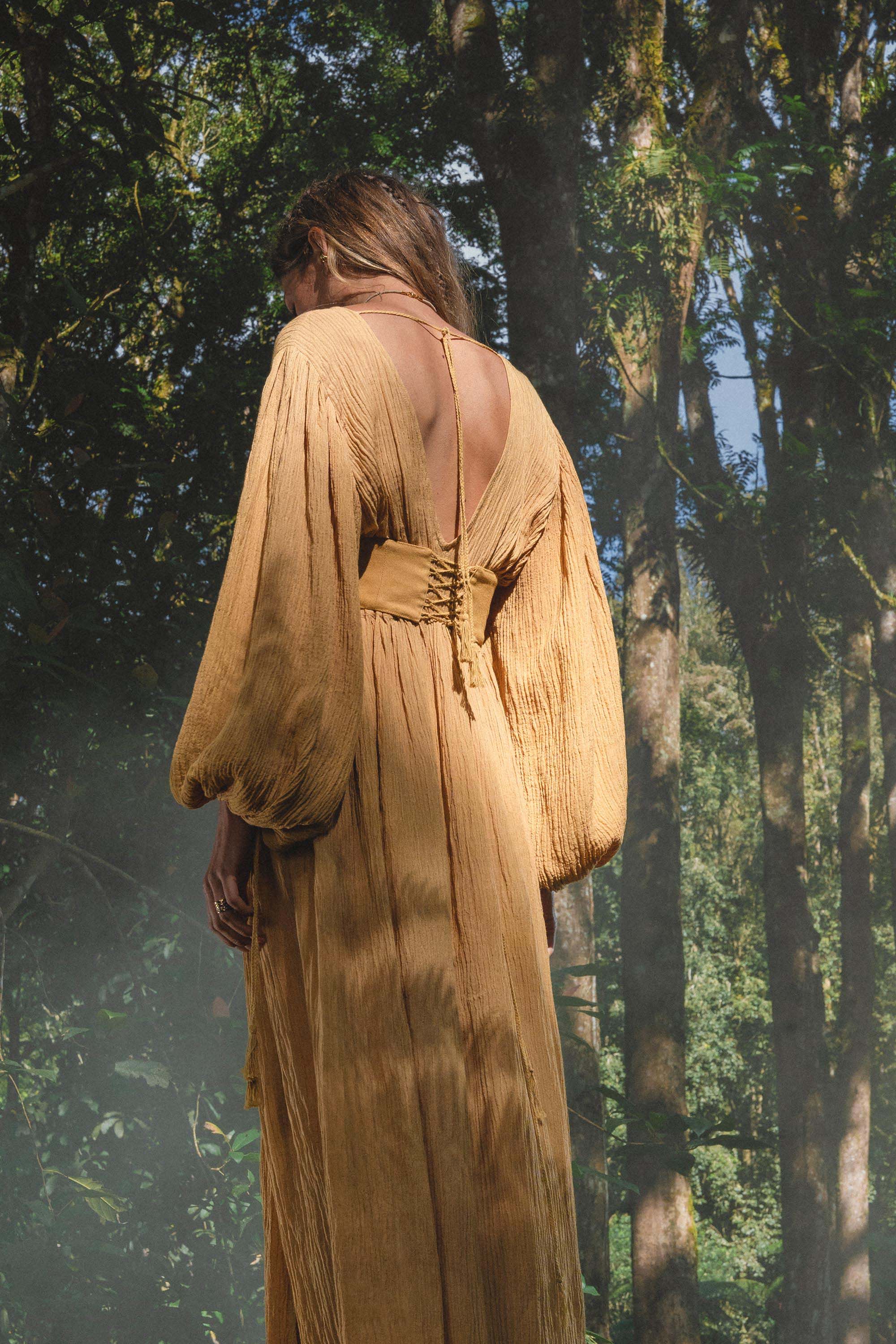 A person stands in a sunlit forest wearing the AYA Sacred Wear Tatva Dress in Saffron Gold—a botanically dyed, organic dress with voluminous sleeves and an open back tied at the neck. Soft light creates a serene, natural setting.