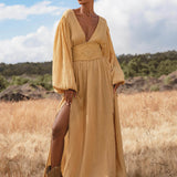 In a dry grassy field, a woman wears the AYA Sacred Wear Tatva Dress Saffron Gold Colour – Organic Goddess Dresses, featuring billowy sleeves and a deep V-neck with a high slit. She stands confidently beneath a partly cloudy sky, trees in the background.
