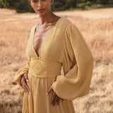 A woman stands in a golden field wearing the Tatva Dress Saffron Gold Colour by AYA Sacred Wear—a long, botanically dyed organic dress with wide sleeves, deep V-neck, and handcrafted belt—gazing softly at the camera amid trees and blue sky.