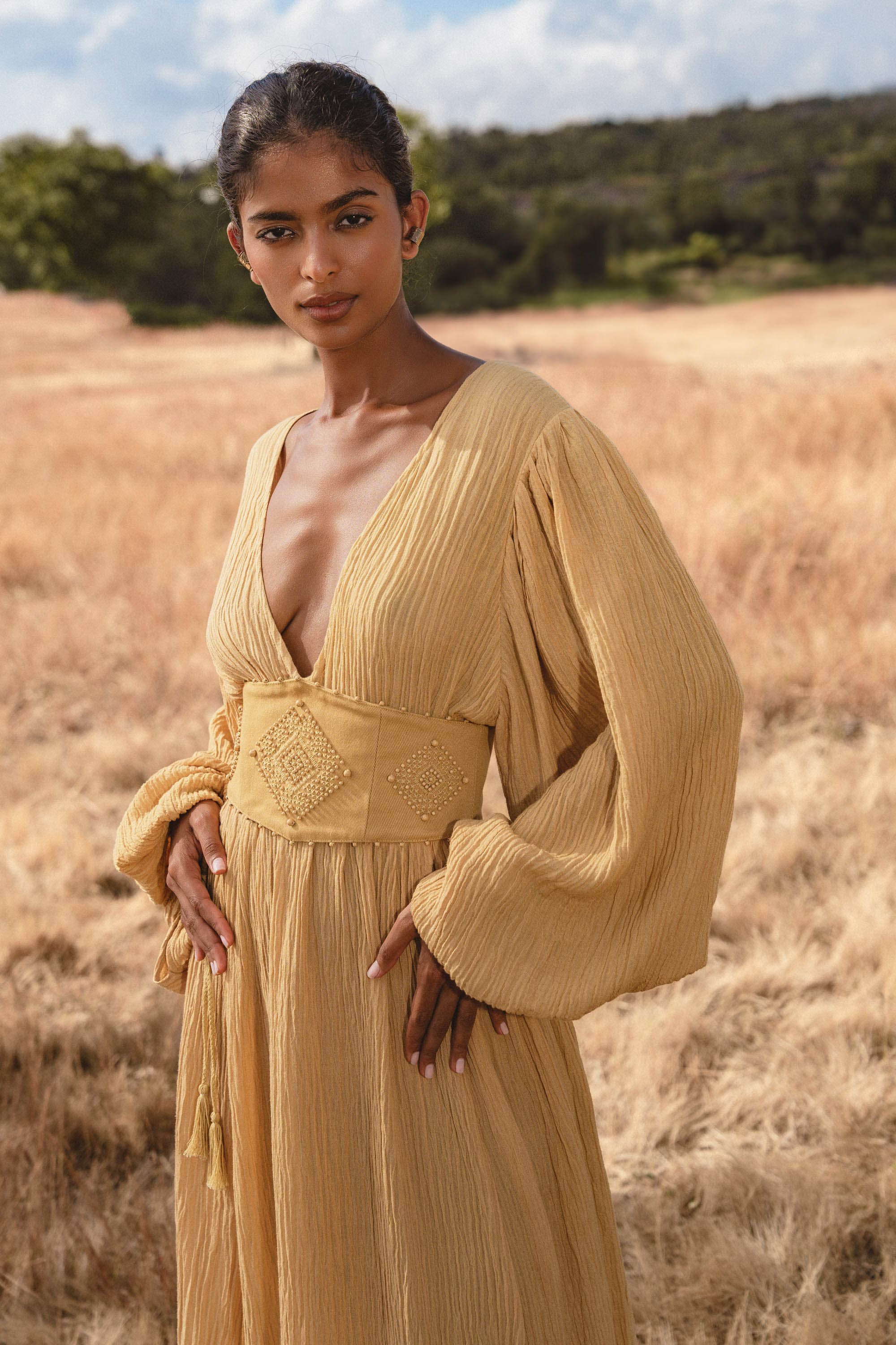 A woman stands in a golden field wearing the Tatva Dress Saffron Gold Colour by AYA Sacred Wear—a long, botanically dyed organic dress with wide sleeves, deep V-neck, and handcrafted belt—gazing softly at the camera amid trees and blue sky.