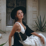 A woman sits gracefully outside, her organic cotton white dress complemented by an AYA Sacred Wear Under Bust Corset for Women, adorned with a wide-brimmed hat. She is poised against a rustic backdrop of plants and a wooden door, her serious expression adding an elegant and enigmatic touch to the scene.