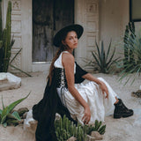 A woman in an under-bust corset from AYA Sacred Wear, part of their boho bridal collection, sits on a sandy terrain amidst cacti. She accessorizes her look with a wide-brimmed black hat and matching boots, while a black shawl is elegantly draped over her shoulder. In the backdrop, there stands a rustic thatched-roof building.