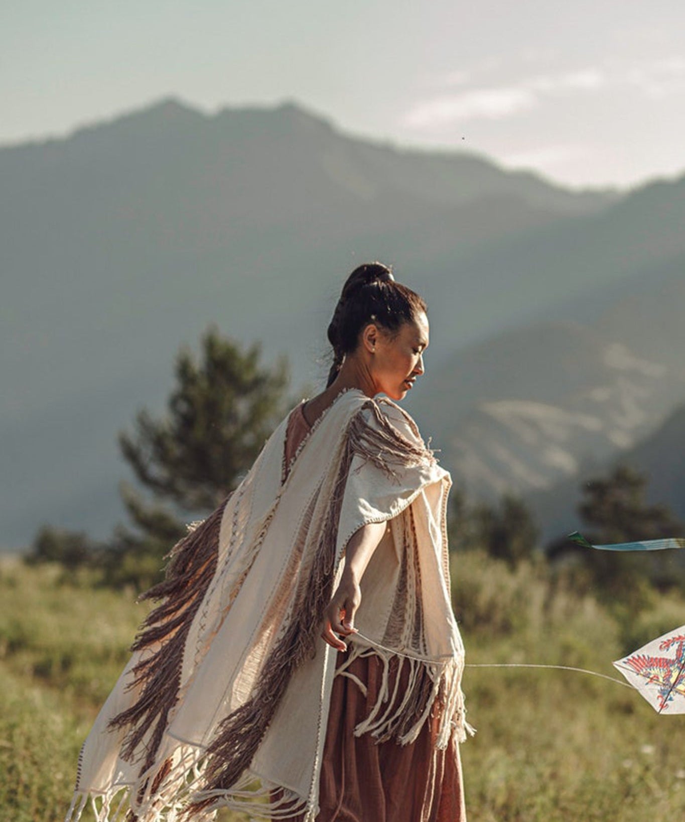Wearing the "Unique Off-White Boho Poncho with Hand Loomed Tassels" by AYA Sacred Wear, a person gracefully flies a kite within a mountainous landscape. Facing to the right, they are surrounded by sunlit mountains and trees, forming a serene and picturesque scene.