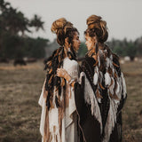 Two people stand in a field, facing each other with arms gently resting on each other's shoulders. They are dressed in bohemian-style clothing, including the Unique Off-White Boho Poncho with Hand Loomed Tassels by AYA Sacred Wear. The background features trees and distant animals under a grey sky.
