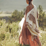 In a lush, sunlit field, a woman stands wearing a flowing pink dress paired with AYA Sacred Wear's Unique Off-White Boho Poncho featuring hand loomed tassels. Her hair is styled in a loose bun adorned with decorative feathers as she gazes into the distance, enveloped by greenery and soft, natural light.