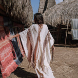 A person wearing the AYA Sacred Wear's Unique Off-White Boho Poncho with Hand Loomed Tassels strolls through a village filled with traditional thatched huts, admiring the hand embroidery on colorful woven textiles. The bright, sunlit scene highlights the intricate artistry and craftsmanship.