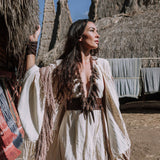 A woman stands outdoors in a Unique Off-White Boho Poncho with Hand Loomed Tassels by AYA Sacred Wear, adorned with bead necklaces. She gazes to the side amidst tall, thatched structures and colorful textiles hanging nearby under a clear blue sky.