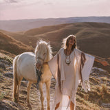 A person stands on a scenic path with a white horse, adorned in an elegant flowing dress layered with AYA Sacred Wear's Unique Off-White Boho Poncho, featuring delicate handloomed tassels and intricate embroidery. The landscape of rolling hills is bathed in warm, golden light, creating a serene and picturesque atmosphere.
