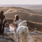 Two people in flowing garments walk hand in hand with a white horse across a rocky hillside at sunset, one of them elegantly draped in AYA Sacred Wear's Unique Off-White Boho Poncho featuring Hand Loomed Tassels. Rolling hills stretch out in the background under a soft, dimming sky.