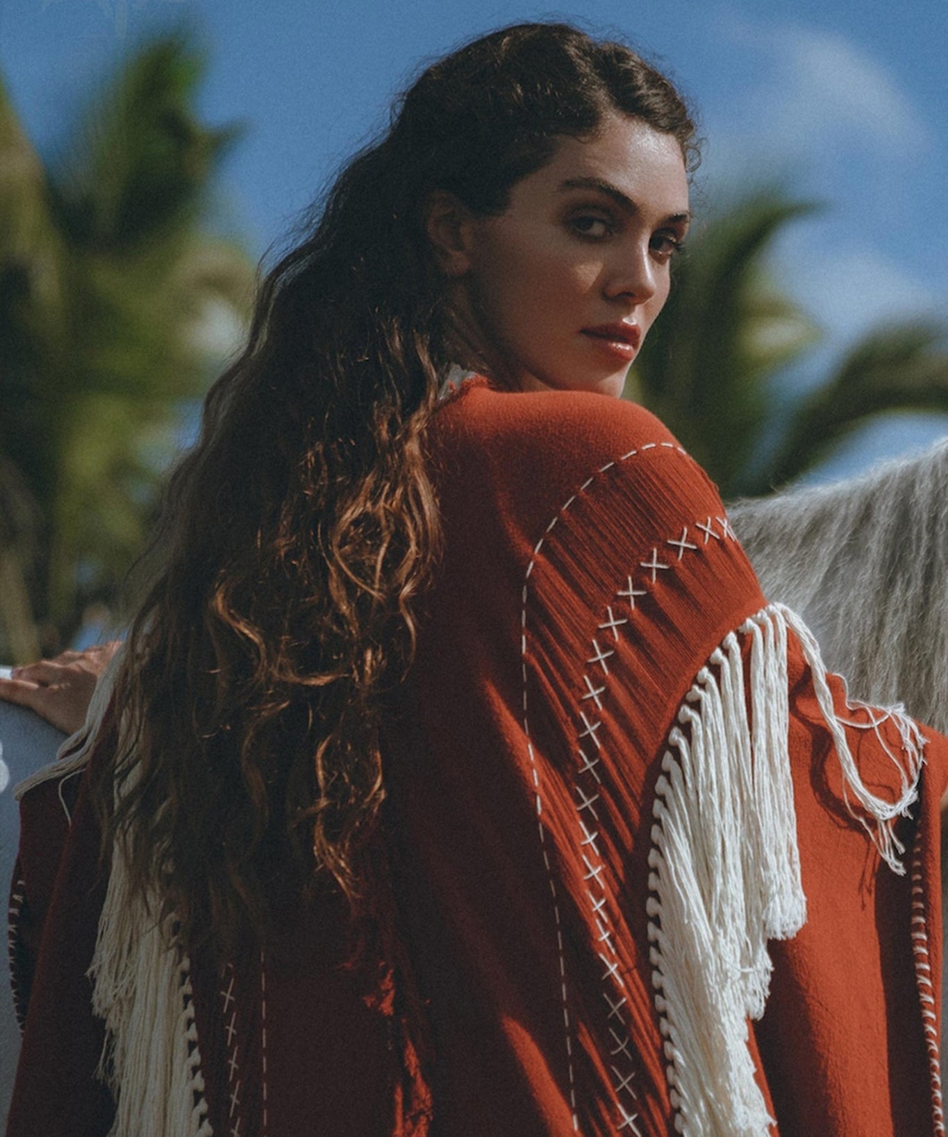 A woman with long, wavy hair stands outside wearing an Unisex Handwoven Cotton Wine Red Poncho from AYA Sacred Wear, featuring a white fringe. She glances back over her shoulder. In the background, palm trees and a blue sky with a few clouds are visible.