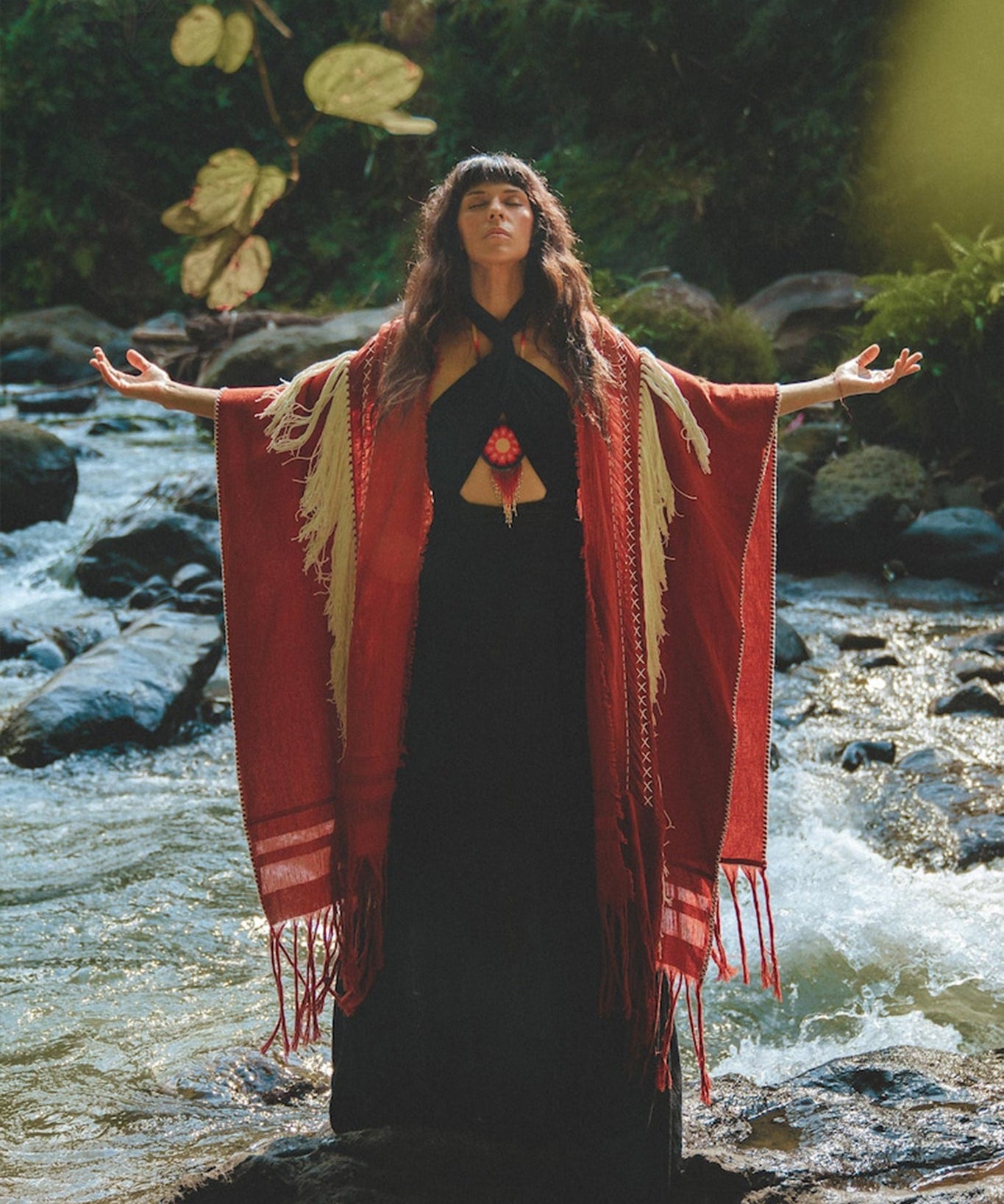 A woman stands on rocks in a flowing stream, arms outstretched. She wears an AYA Sacred Wear Unisex Handwoven Cotton Wine Red Poncho over a black dress. The lush, green setting with sunlight filtering through the trees creates a serene, nature-focused scene.