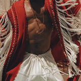 A person dances energetically, draped in an Unisex Handwoven Cotton Wine Red Poncho from AYA Sacred Wear, paired with white pants. The poncho sways dynamically, capturing motion and joy against a backdrop of intricate decorative patterns.