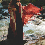 A woman stands gracefully by a flowing stream, enveloped in an AYA Sacred Wear Unisex Handwoven Cotton Wine Red Poncho over her long black dress. She gazes downward, surrounded by lush greenery in a serene, natural setting.