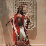 Wrapped in an AYA Sacred Wear Unisex Handwoven Cotton Wine Red Poncho and white pants, a person poses dramatically by a turquoise pool. Behind them, intricate white wall art creates a stunning backdrop as shadows of palm leaves enhance the shamanic tropical ambiance of the boho scene.