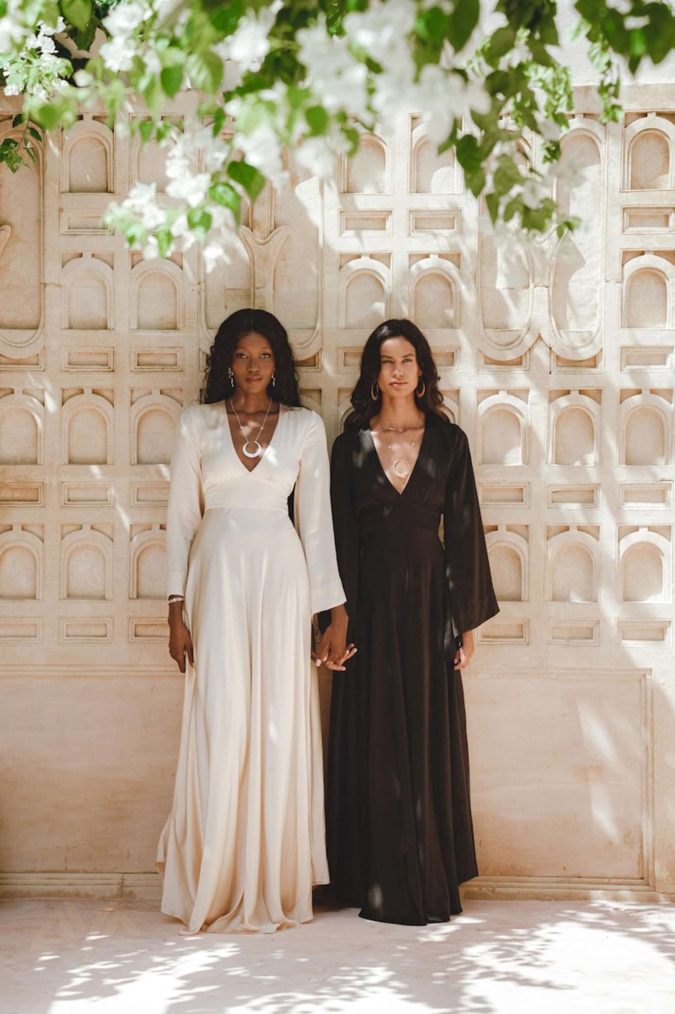 Two women stand side by side against an ornate wall. One wears AYA Sacred Wear's Vanilla Cream Sundari Silk Gown, while the other is in a long black dress. Sunlight creates patterns of light and shadow as they wear jewelry and hold hands in serene unity.