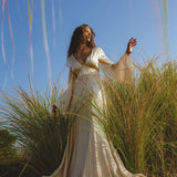A woman in a Vanilla Cream Sundari Silk Gown by AYA Sacred Wear stands amid tall grass under a clear sky, extending her arm gracefully and gazing ahead, creating an elegant and ethereal scene.