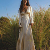 Amidst tall grass, a woman with long curly hair stands wearing the elegant Vanilla Cream Sundari Silk Gown by AYA Sacred Wear. The clear blue sky hints at a sunny day, creating a serene, luxurious atmosphere.