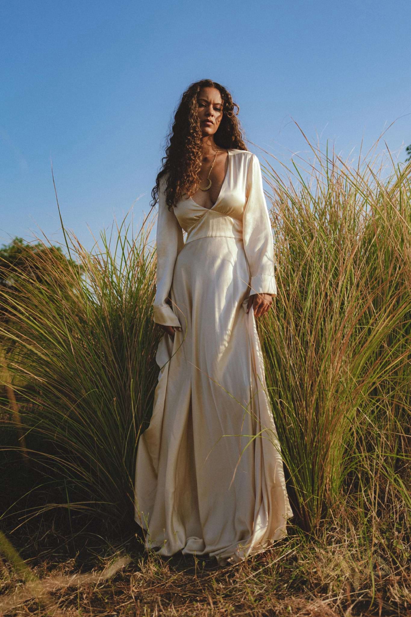 Amidst tall grass, a woman with long curly hair stands wearing the elegant Vanilla Cream Sundari Silk Gown by AYA Sacred Wear. The clear blue sky hints at a sunny day, creating a serene, luxurious atmosphere.