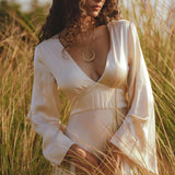 A woman with long curly hair stands in tall grass, wearing an AYA Sacred Wear Vanilla Cream Sundari Silk Gown and a crescent moon necklace. She gazes downward thoughtfully, bathed in warm sunlight against a softly blurred green background.