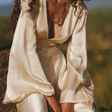 A person with long, curly hair sits outdoors on a rock wearing an AYA Sacred Wear Vanilla Cream Sundari Silk Gown with a deep V-neckline. A pendant necklace adorns their neck, surrounded by blurred greenery in the background.