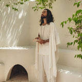 A person with long hair stands barefoot in a serene, sunlit courtyard, surrounded by leafy green branches. They are wearing an AYA Sacred Wear White Raw Cotton Minimalist Poncho for Men and holding a book, exuding a peaceful, contemplative presence.
