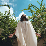 A figure with long hair stands outside, draped in a White Raw Cotton Minimalist Poncho for Men by AYA Sacred Wear. With arms open, they gaze at the bright blue sky. Green foliage and white flowers envelop them, enhancing the tranquil and serene setting. The botanically dyed handwoven cotton of the poncho perfectly harmonizes with the surrounding natural beauty.
