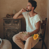 A man with a beard sits thoughtfully in a chair, wearing the White Raw Edge Thick Cotton Top Tee for Men by AYA Sacred Wear and beige pants. A sunflower peeks from his pocket and beads adorn his neck and wrist. The T-shirt, made from handwoven organic cotton, is botanically dyed. Behind him is a rustic wooden cabinet against a neutral-colored wall.